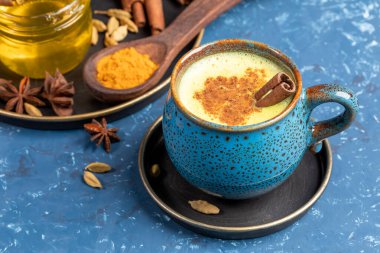 Mavi fincanda geleneksel Hint ayurvediği altın zerdeçal latte sütü. Mavi arka planda kakule, bal ve tarçın çubuğu var. Bağışıklığı geliştirmek için sağlıklı bir içecek. Seçici odak.