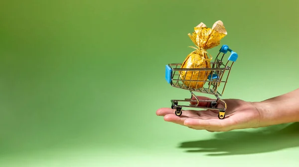 Bayramlık Paskalya bayrağı. Bir adet altın Paskalya yumurtası desenli kağıda sarılmış küçük dekoratif süpermarket sepeti tutan uzanmış kadın eli. Tatil teslimatı konsepti. Metin için alanı kopyala.