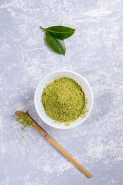 Beyaz kasede yeşil Japon kibrit çay tozu, özel ahşap bambu kaşığı ve gri desenli arka planda taze yapraklar.. 