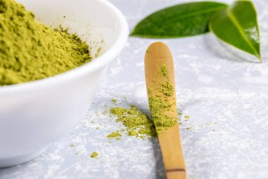 Kapalı ahşap bambu kaşığı, yeşil Japon kibrit çay tozu, çay tozlu beyaz kase ve gri desenli arkaplanda taze yapraklar. Sağlık için antioksidan kaynağı. Seçici odak.