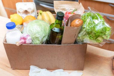 Ahşap mutfak masasında yeni paketlenmiş gıda ürünleri olan karton kutu. Güvenli teslimat konsepti. Sosyal gıda bağışı. Karantina sırasında güvenli ev yemeği teslimatı. Seçici odak.