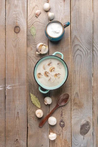 Vertical arrangement of turquoise ceramic pan with mushroom cream soup, cup with sour cream, bowl with fresh champignons, spoon, garlic and bay leafs on wooden background. Copy space.