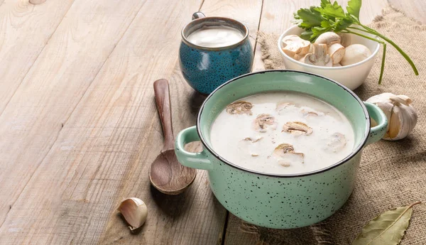 Turquoise ceramic pan with mushroom cream soup, cup with sour cream, bowl with fresh champignons, spoon and spices on burlap on wooden background. Copy space.