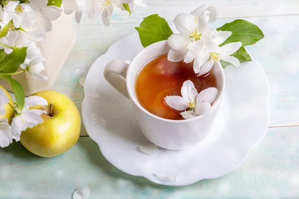 White cup of tea on shaped saucer with blossom apple tree flowers and yellow apple on wooden table with bokeh closeup. — Stock Photo, Image