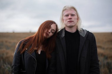 Genç albino ve kızıl saçlı bir kız tarlada dikiliyor, adam kameraya bakıyor.