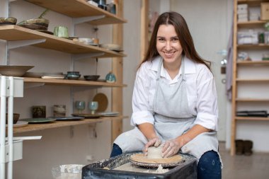 Genç seramikçi çömlekçinin tekerinin arkasında oturur kameraya bakar ve gülümser.