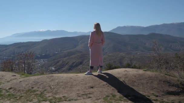 Bir kız dağın tepesinde duruyor ve dağların güzelliğinin tadını çıkarıyor.. — Stok video