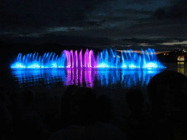 Fountains of blue and crimson at night over the water.