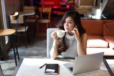 Güzel ve çekici Asyalı kadın, gri günlük elbiseli uzun saçlı sabahları kahve dükkanında bilgisayar kullanırken masada çalışıyor ve kahve içiyor.