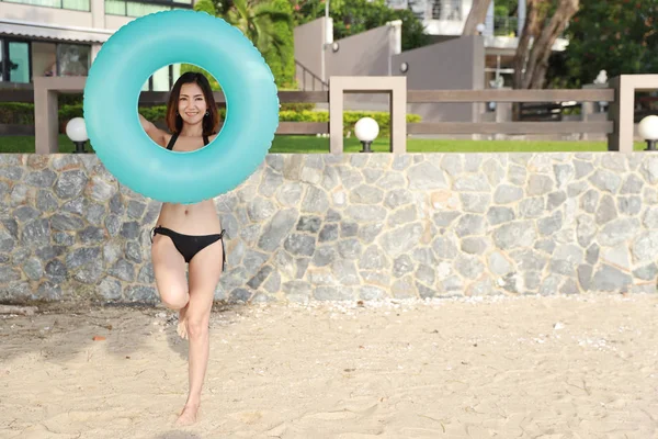 Mutlu, genç ve güzel Asyalı bir kadın, siyah bikinili, mavi şişme bir can simidiyle kumsalda güneşli bir günde gülen yüzüyle eğleniyor. — Stok fotoğraf