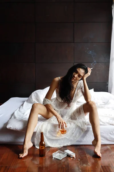 Повна довжина п'яна азіатська жінка в білій білизні, пияцтво і куріння, тримаючи пляшку алкоголю і сидячи на ліжку в спальні — стокове фото