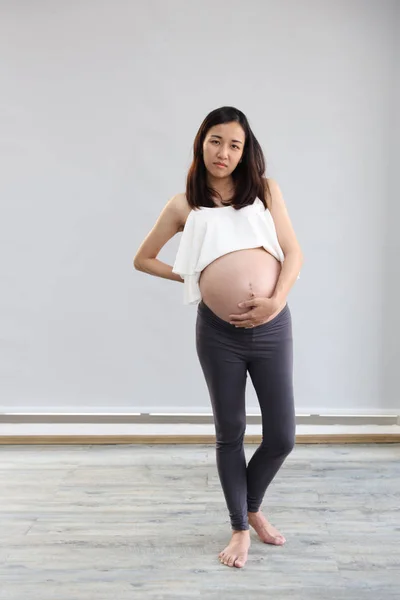 Junge asiatische schwangere Frau mit weißem Kleid und langen Haaren steht und hält ihren Bauch mit besorgtem und unglücklichem Gesicht, während sie morgens in die Kamera auf weißem Hintergrund im Wohnzimmer schaut — Stockfoto