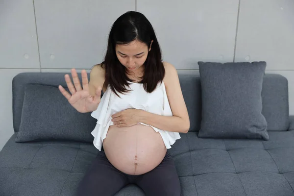 Mladý asijské těhotná dívka v bílých šatech s dlouhými vlasy sedí na pohovce v obývacím pokoji. Roztomilá žena zvedne ruku k někomu, kdo chce něco odmítnout (těhotné a zdravotní péče) — Stock fotografie