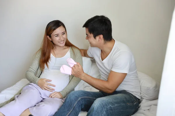 Nahaufnahme asiatische schwangere Frau und kaukasischer Ehemann sitzen auf dem Bett und halten rosa Babysocken, während die Augen Kontakt miteinander haben. — Stockfoto