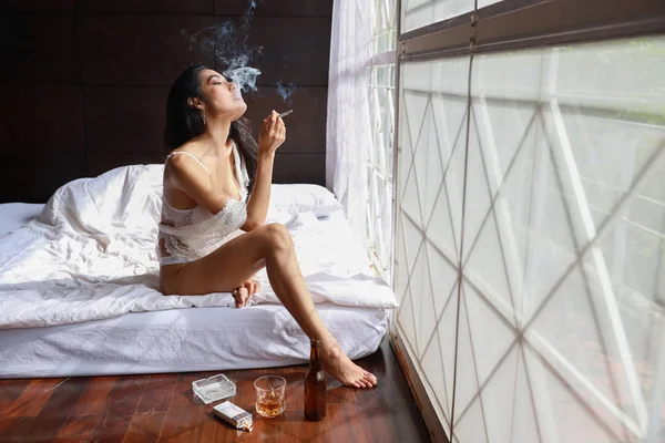 アジア系の酔った女性が白いランジェリーを着て酒を飲みタバコを吸いながらベッドルームで酒を飲んで — ストック写真