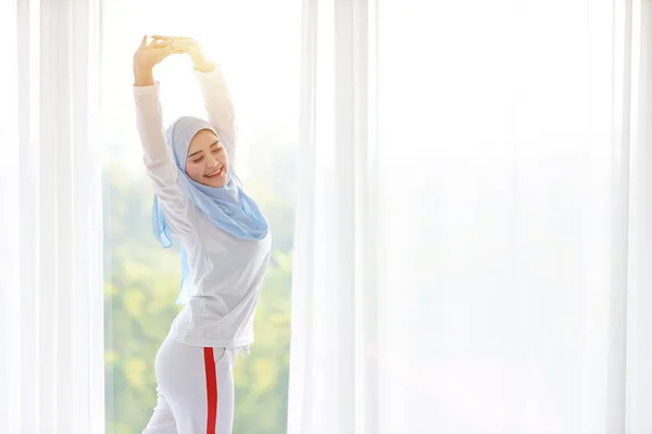 Wanita muslim Asia yang cantik mengenakan pakaian tidur putih, meregangkan lengannya setelah bangun di pagi hari saat matahari terbit. Gadis muda yang lucu dengan hijab biru berdiri dan bersantai dengan wajah gembira dan tersenyum. — Stok Foto