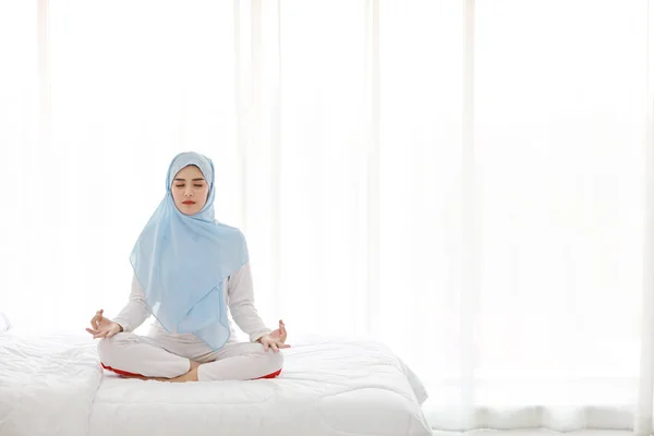若いアジアのイスラム教徒の女性がベッドに座って瞑想を楽しんでいます。青のヒジャーブの練習ヨガと寝室での美しい女の子は、平和と穏やかな。健康的でライフスタイルのコンセプト — ストック写真