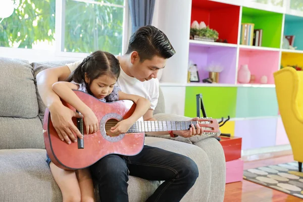 Szczęśliwy azjatycki tata uczy jego córka grać na gitarze akustycznej na szarej kanapie w salonie ze śmiechem i uśmiechem na twarzy (koncepcja stylu życia) — Zdjęcie stockowe