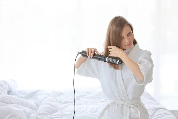 Giovane donna caucasica bella in pigiama da notte seduta sul letto e utilizzando piastra per capelli e sorridente in camera da letto dopo il risveglio al mattino — Foto Stock