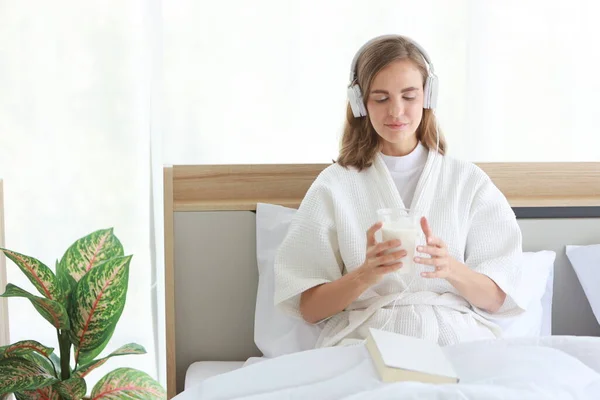 Jovem mulher caucasiana alegre em pijama branco com fone de ouvido que segurando um copo de leite enquanto lê um livro antes de ir dormir com o rosto feliz e sorriso na cama no quarto moderno branco . — Fotografia de Stock