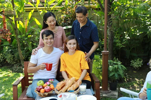 Happy multiethnic family sitting at a breakfast table in backyard outdoor on sunny day with smiling face. Happy caucasian and asian family sitting and having conversation with good time outdoor. — Stock Photo, Image