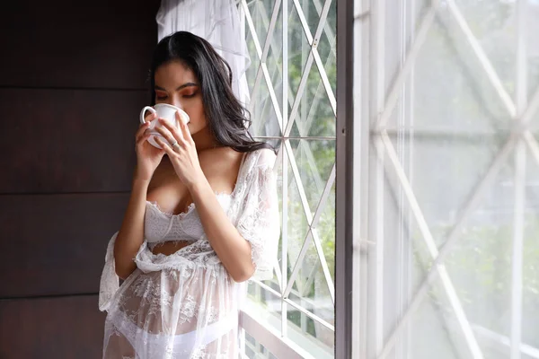 Retrato de mulher asiática atraente e sexy em roupa de dormir lingerie branca em pé ao lado da janela e beber café pela manhã com relaxamento. Jovem bonito menina longo cabelo em pé no quarto — Fotografia de Stock
