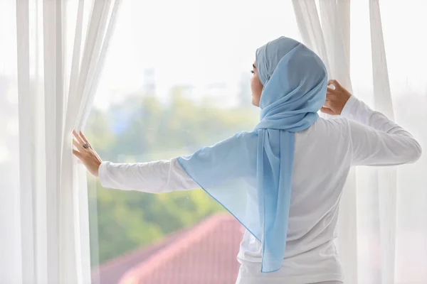 Rear view beautiful asian muslim woman wearing white sleepwear, stretching her arms after getting up in the morning at sunrise. Cute young girl with blue hijab standing and relaxing while looking away — Stock Photo, Image
