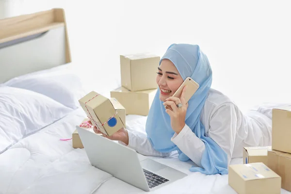 Gyönyörű és fiatal ázsiai muszlim nő hálóingben vonzó megjelenés, fekszik az ágyban a számítógép és az online csomag doboz szállítás. Induló kisvállalkozás KKV szabadúszó, mobiltelefonnal dolgozó lány — Stock Fotó