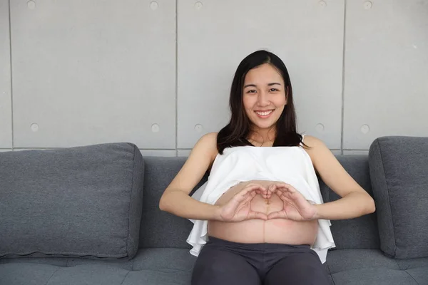 Hermosa y joven mujer embarazada asiática en camisa blanca haciendo símbolo del corazón en su vientre y sentada en un sofá gris en la sala de estar con la cara feliz y sonriendo. Publicidad retrato embarazo concepto —  Fotos de Stock
