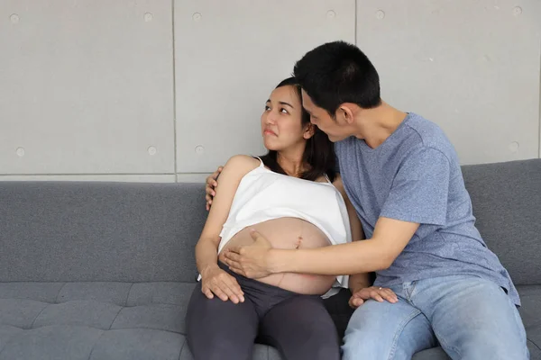 Junger asiatischer Ehemann in blauem T-Shirt tröstend, umarmt seine junge unglückliche asiatische schwangere Frau, während er mit lächelndem Gesicht auf dem Sofa im Wohnzimmer sitzt. Werbeporträt Schwangerschaftskonzept — Stockfoto