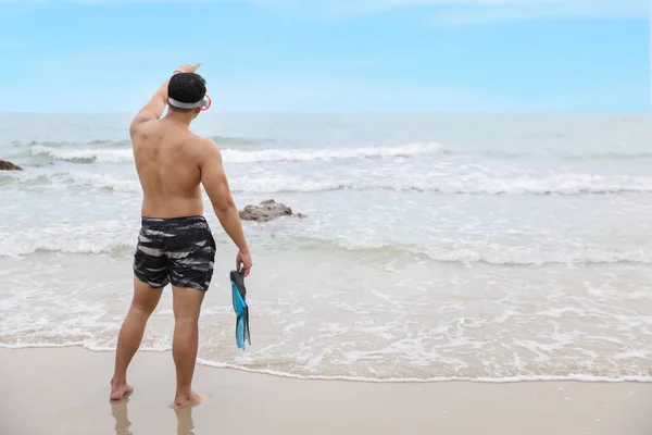 Voltar vista retrato de jovem asiático bonito e sexy homem em preto natação terno, ele segurando azul mergulhador e vestindo snorkel enquanto em pé e apontando algo na praia tropical no verão — Fotografia de Stock