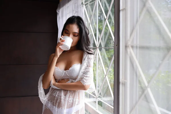 Retrato de mulher asiática atraente e sexy em roupa de dormir lingerie branca em pé ao lado da janela e beber café pela manhã com relaxamento. Jovem bonito menina longo cabelo em pé no quarto — Fotografia de Stock