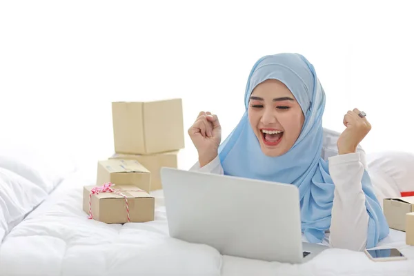 Gyönyörű és fiatal ázsiai muszlim nő hálóingben vonzó megjelenés, fekszik az ágyban számítógép, mobil telefon és online csomag doboz szállítás. Okos lány hidzsáb kap jó hír és meglepetés. — Stock Fotó