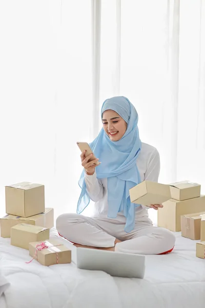Active smiling asian muslim woman in sleepwear sitting on bed using mobile phone and computer. Startup small business SME freelance girl working with online package box delivery, E-commerce concept. — Stock Photo, Image