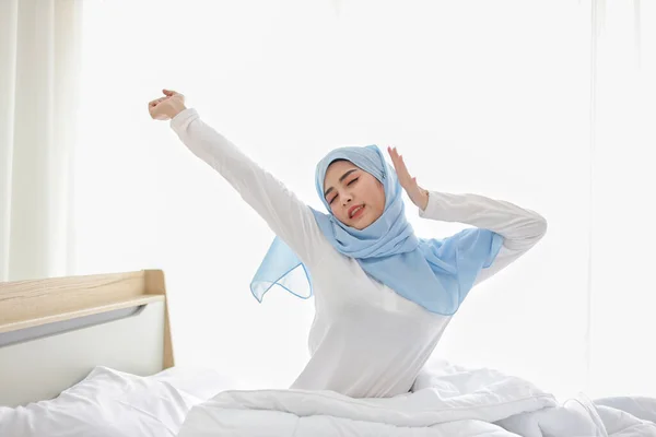 Beautiful asian muslim woman wearing white sleepwear sitting on bed, stretching her arms after getting up in the morning at sunrise. Cute young girl with hijab wake up and relaxing, closing her eyes — Stock Photo, Image