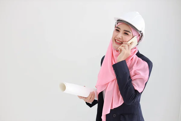 活跃而聪明的穆斯林年轻的亚洲女人穿着蓝色西服，微笑着，自信地拿着蓝图，在工作室里使用手机。与工程系女孩隔离的白色背景。广告概念. — 图库照片