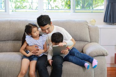 Mutlu Asyalı baba, kızına oturma odasındaki gri koltukta gülen ve gülen yüzlü tablet kullanmayı öğretiyor.)