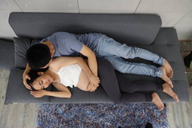 Genç Asyalı koca kanepeye uzanıp hamile karısına sevgi ve mutlulukla sarılıyor ve onu öpüyor.