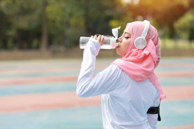 Spor yaptıktan sonra su içen güzel fitness sporcusu Asyalı Müslüman kadın. Spor kıyafetli, kulaklıklı, dışarıda antremandan sonra dinlenen hoş bir kız. Sağlıklı, spor konsepti