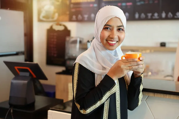 迷人的穆斯林女人年轻的亚洲女人站在咖啡店的柜台边 一边站着一边喝咖啡 一边笑着看着相机 — 图库照片