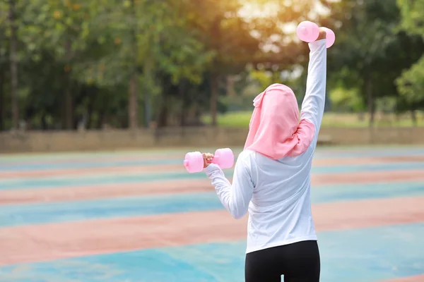 Spor giyim kuşağında genç Asyalı atletik Müslüman kadın sabah egzersizi için ayakta duruyor ve dambılları kaldırıyor. Aktif kız, ağaç köklü pazı egzersizi yapıyor. Spor konsepti