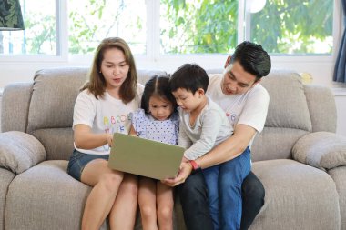Mutlu Asyalı aile çocuklarına bilgisayar kullanmayı öğretirken oturma odasındaki gri koltukta gülen yüzlerle oturuyorlar.
