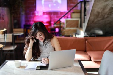 Güzel ve çekici Asyalı kadın, kafeteryada cep telefonu ve dizüstü bilgisayar kullanırken evrak işleriyle uğraşan gri günlük elbiseli uzun saçlı.