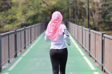 Arka manzara portresi sporcu genç Asyalı müslüman kadın spor giyim maraton antrenmanı için açık havada koşuyor. Yeşil ağaç zeminli, beton yol boyunca egzersiz yapan koşucu kız. Spor konsepti