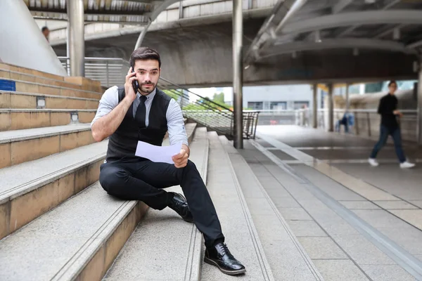 successful caucasian businessman in black suite who sitting in downtown, he using cell phone and looking away while holding paper work with city background