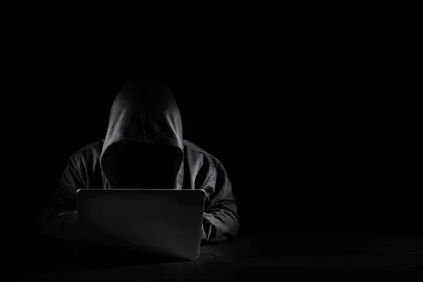 Dangerous anonymous hacker man in black hooded using computer, breaking into security data corporate server. He sitting, working on black background. Internet crime, cyber attack security concept