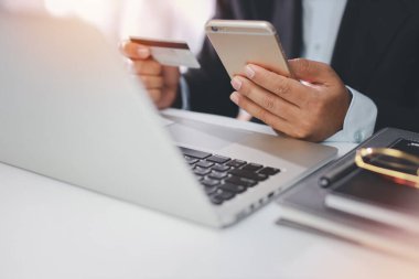 Siyah takım elbiseli bir adam oturuyor, elinde kredi kartı tutuyor ve online ödeme ya da alışveriş için dizüstü bilgisayar kullanıyor. İş adamı çalışırken cep telefonu kullanıyor. E-Bankacılık kavramı