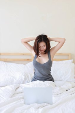 Güzel ve çarpıcı Asyalı serbest çalışan seksi gri elbiseli kadın, ara verip bilgisayar kullanırken yatakta geriniyor. 