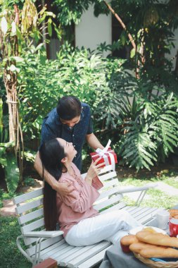 Mavi ve pembe tişörtlü Asyalı yaşlı çift evlilik yıldönümünde arka bahçedeki kahvaltı masasında oturuyor. Asyalı yaşlı koca, gülümseyen yüzlü Asyalı yaşlı karısına sürpriz hediyeler veriyor..