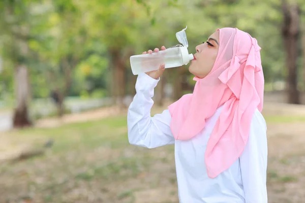 健美的健美运动员亚洲穆斯林妇女在运动后喝水 年轻可爱的女孩穿着运动服 头戴耳机 在户外运动后休息 体育概念 — 图库照片